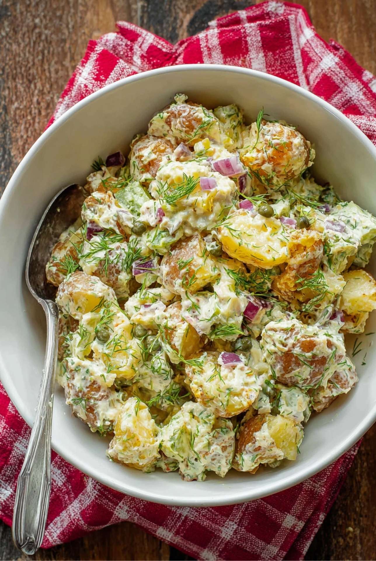 Bowl of smashed potato salad with dill, pickles, and red onion on a wooden table with a red checkered napkin.