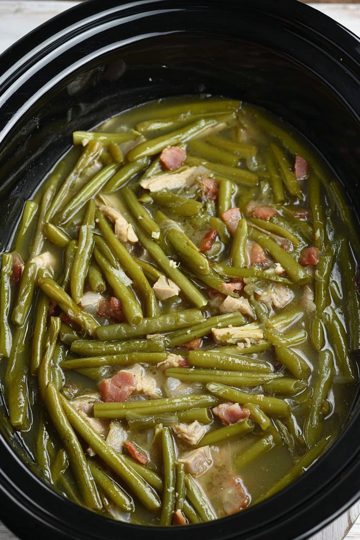 Overhead view of green beans with bacon in a slow cooker, showing tender beans in a savory broth.