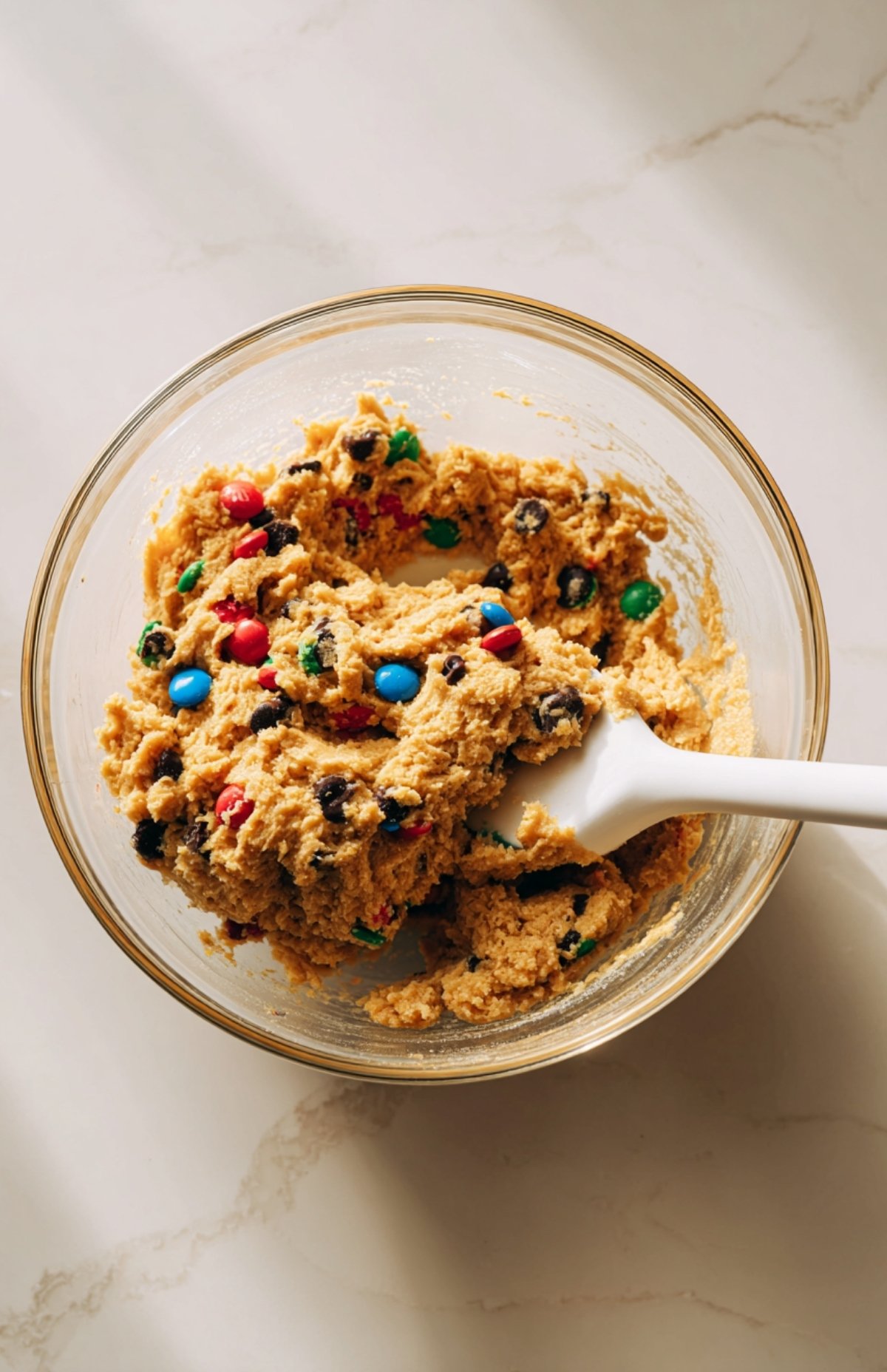 Bowl of cookie dough-style mixture with chocolate chips and colorful M&Ms being folded in with a spatula.
