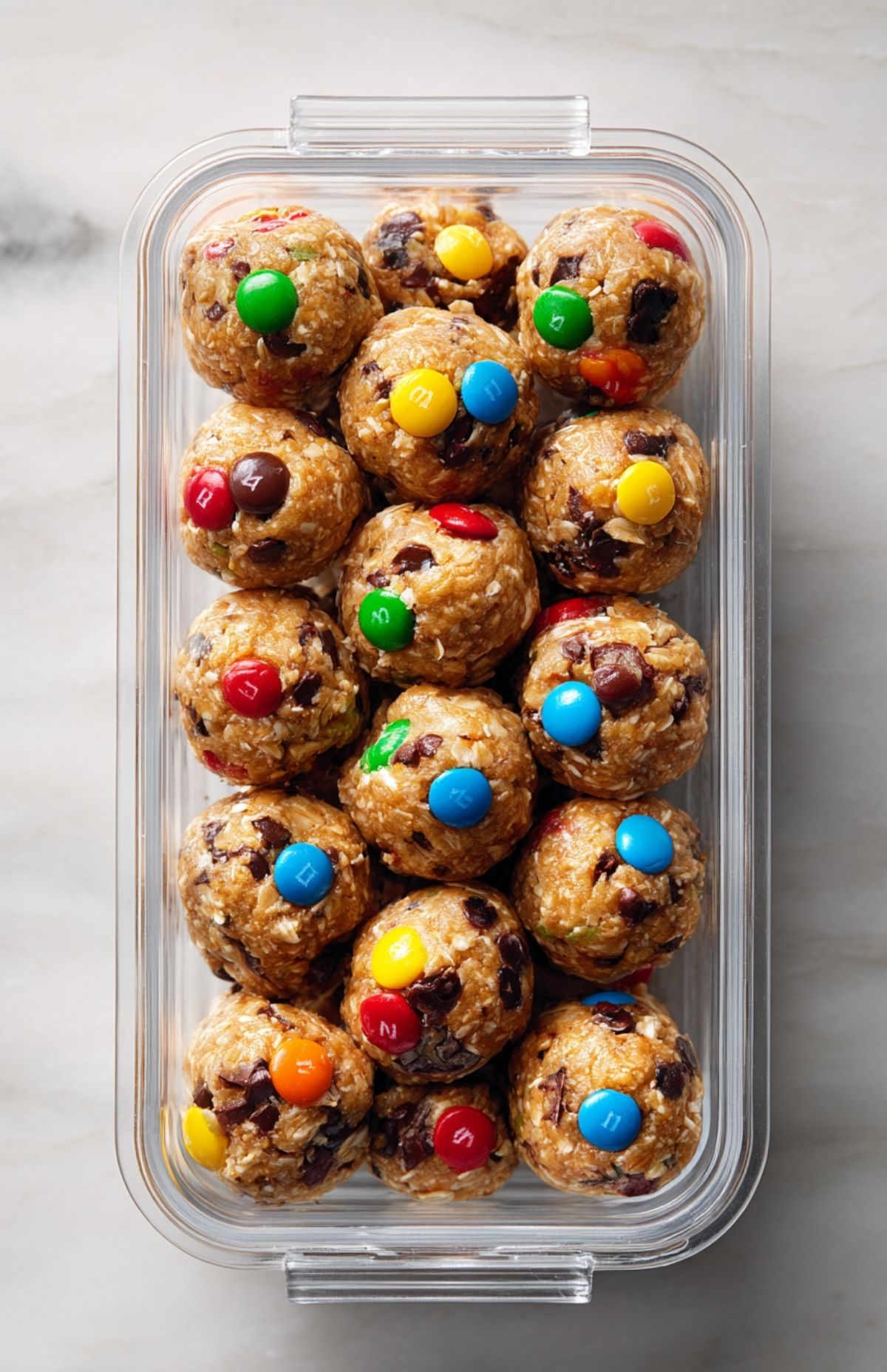 Hands rolling cookie dough mixture into colorful M&M cookie protein balls on a marble counter.