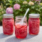 Three glass mason jars packed with sliced pink pickled red onions sitting on a table with pink hydrangeas blurred in the background.