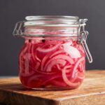 Sliced red onions pickling inside a glass jar with a wire bale latch lid sitting on a wooden cutting board against a dark background.