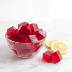 Red gelatin cubes stacked in a clear glass bowl next to fresh lemon slices on a white marble countertop