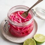 A glass jar filled with bright pink pickled red onions sits on a small speckled plate with two lime halves next to it