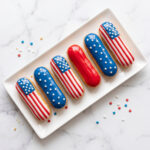 Red, white, and blue decorated éclairs arranged on a white rectangular plate over a marble background.