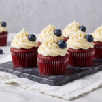 A close-up shot of several Patriotic Red Velvet Cupcakes with swirls of white cream cheese frosting and fresh blueberries on a marble tray.