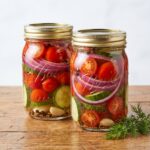 Two glass mason jars filled with layers of halved cherry tomatoes, sliced cucumbers, red onions, garlic cloves, and spices sitting on a rustic wooden table next to fresh dill.