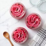 Top down view of three round glass jars packed with vibrant pink pickled red onions and peppercorns on a marble surface next to a wooden spoon.