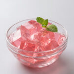A clear glass bowl piled high with pale pink jello cubes and topped with a fresh green mint sprig against a plain gray background.