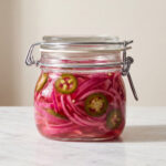 A sealed glass clamp jar filled with bright pink pickled red onions and sliced jalapeños sitting on a white marble tabletop