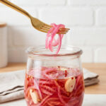 A gold fork lifts thin strands of vibrant pink pickled red onions from a clear glass jar filled with brine, garlic cloves, and red pepper flakes