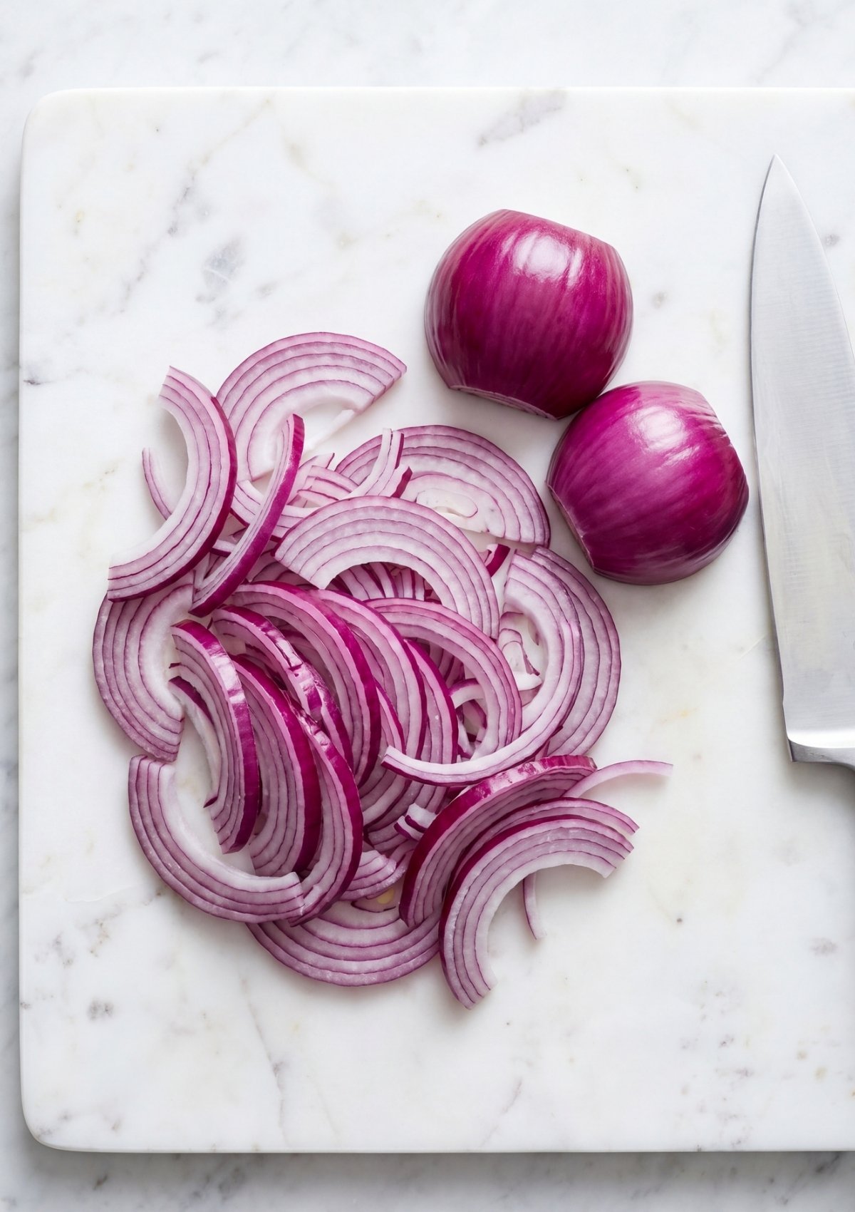 Thinly sliced red onion half rings grouped on a white marble cutting board next to two peeled onion halves and a chef knife