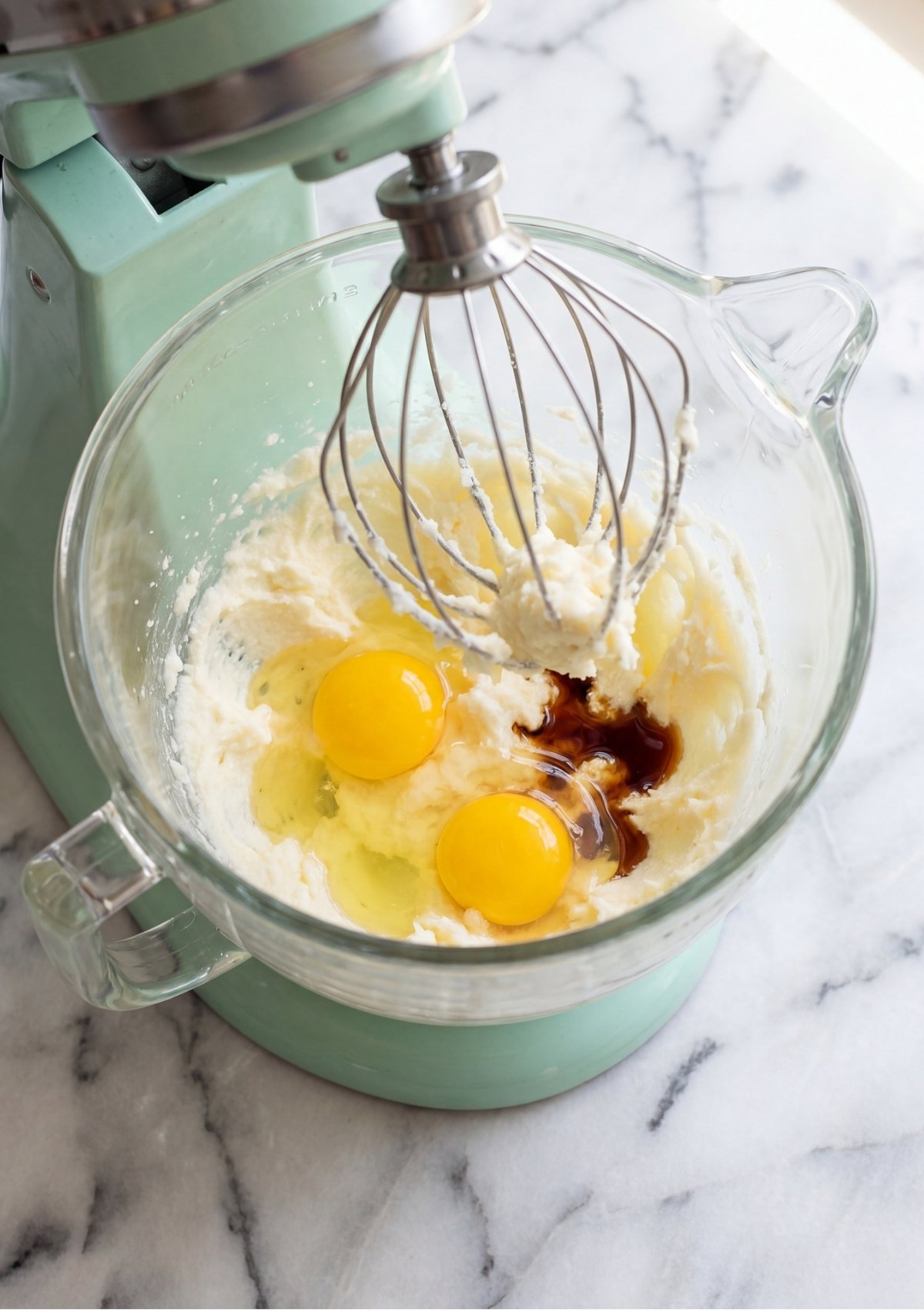 Two bright yellow egg yolks and dark vanilla extract being added to creamed butter and sugar in a glass stand mixer bowl.