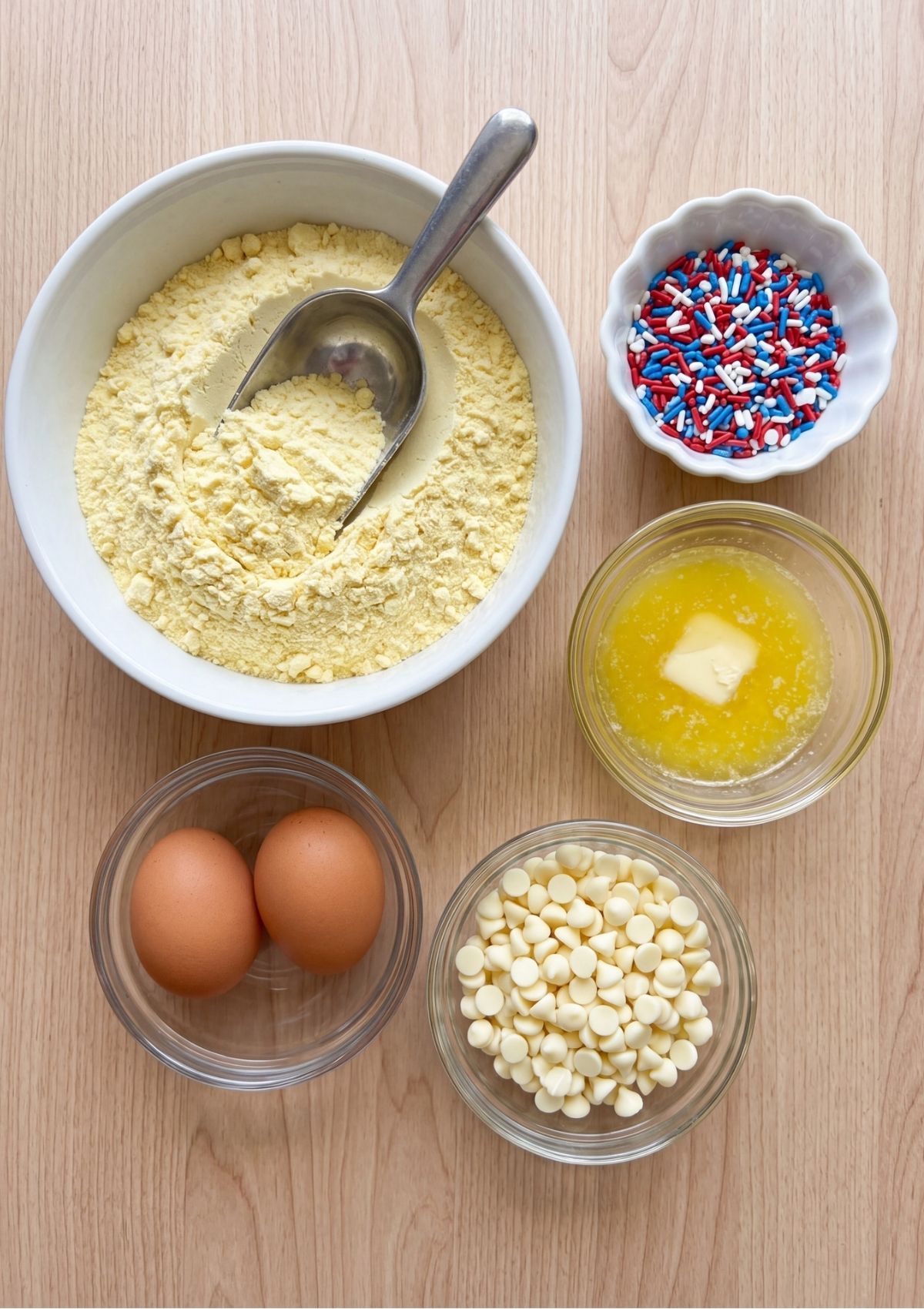 Overhead view of baking ingredients on a wooden surface, including a bowl of yellow cake mix, red, white, and blue sprinkles, melted butter, two eggs, and white chocolate chips.