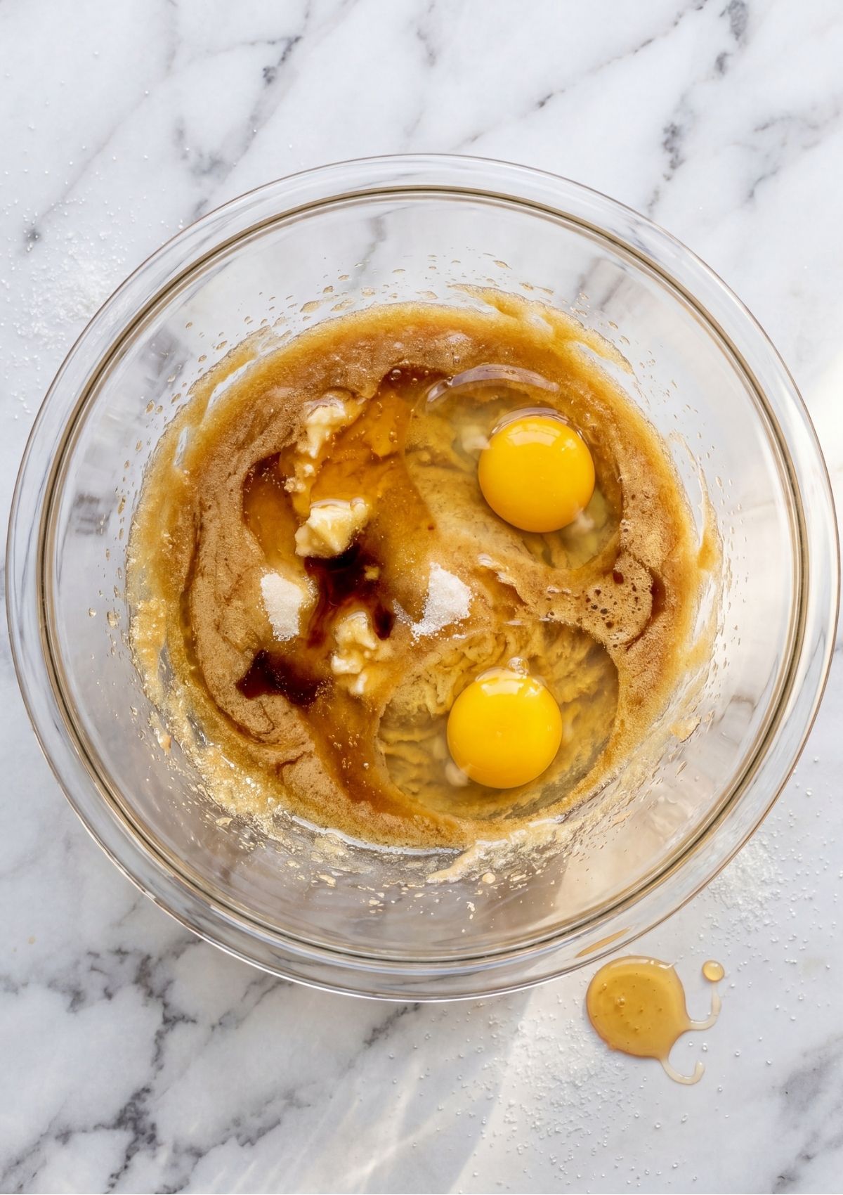 A glass bowl containing cracked eggs, vanilla extract, and melted butter being mixed together.