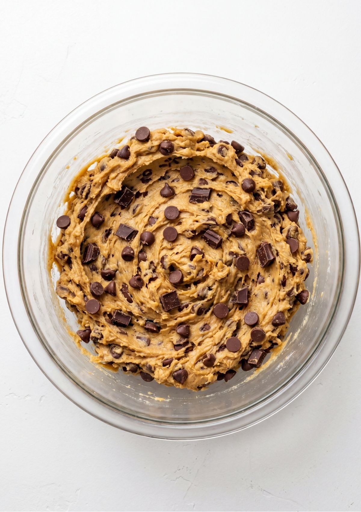A clear glass mixing bowl filled with thick chocolate chip cookie dough topped with chocolate chips and chocolate chunks