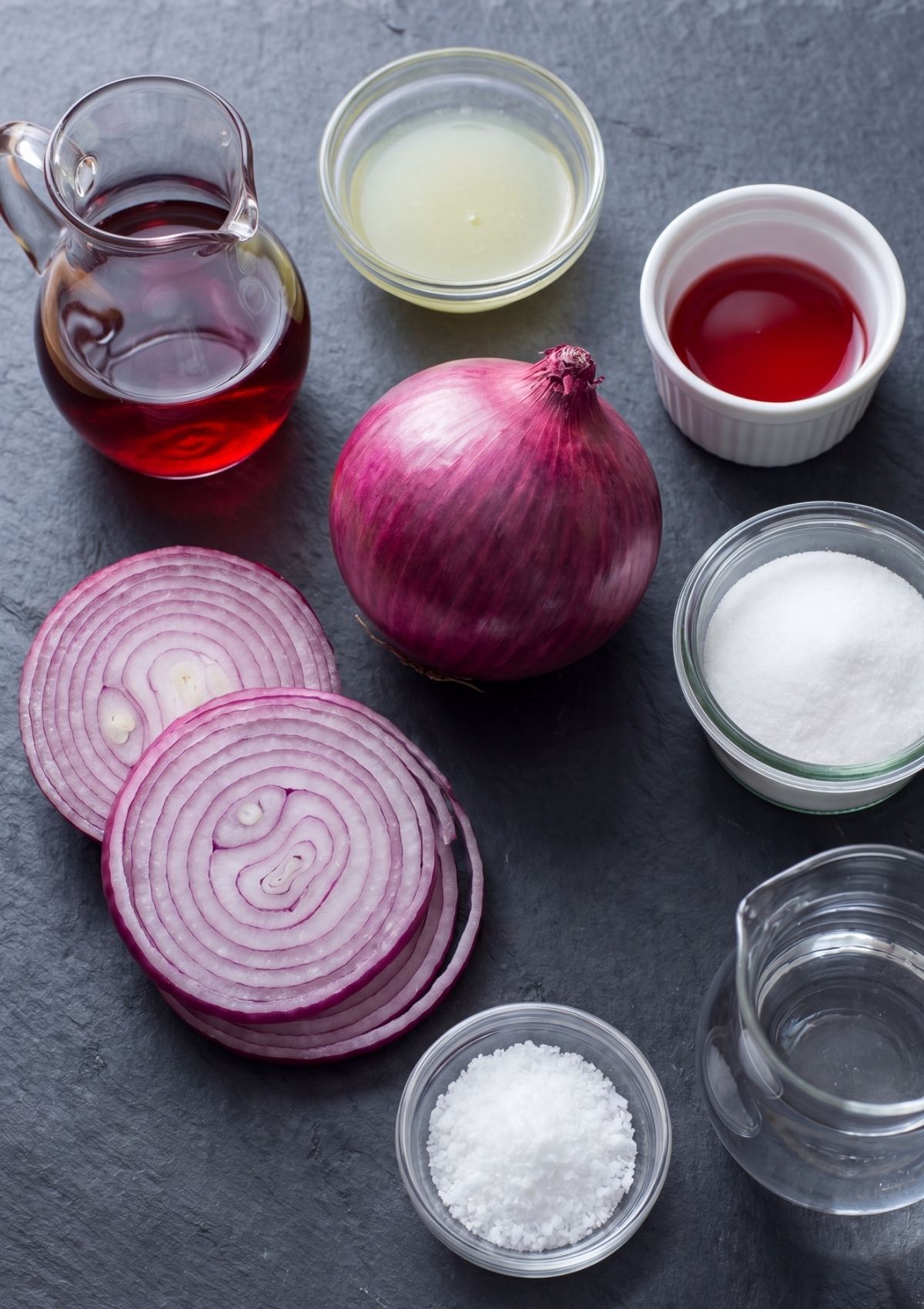 Flat lay of ingredients on a dark slate surface including a whole red onion, sliced onion rings, small bowls of white sugar, coarse salt, red wine vinegar, and fresh lime juice.
