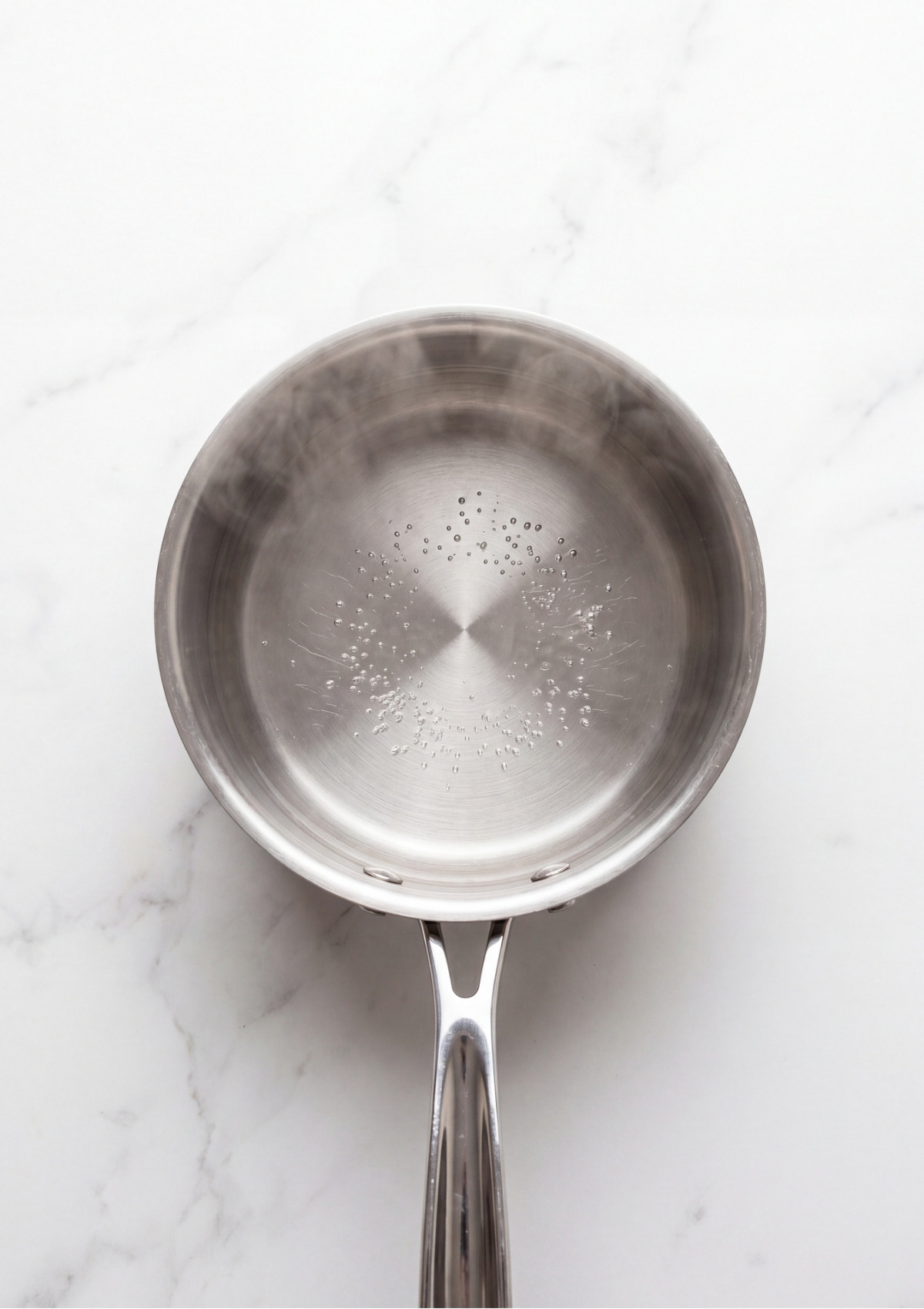 An overhead view of a stainless steel saucepan filled with steaming, hot water with small bubbles forming on the bottom.