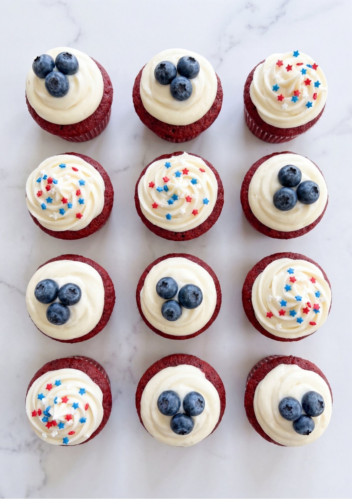 Top-down view of twelve red velvet cupcakes, some topped with three blueberries and others with red, white, and blue star sprinkles.