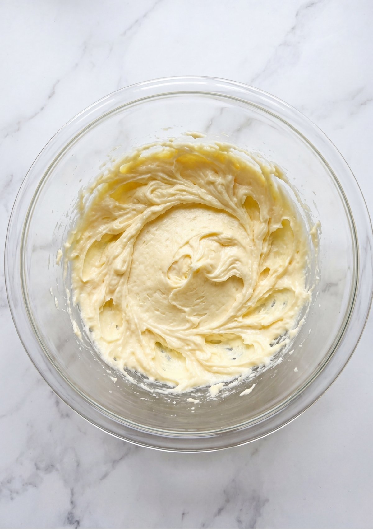 A glass mixing bowl containing a smooth, pale yellow mixture of creamed butter and sugar for the cupcake base.