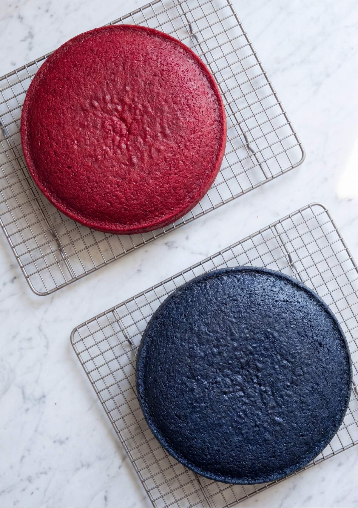 One red cake layer and one blue cake layer cooling on metal wire racks over a white marble background.