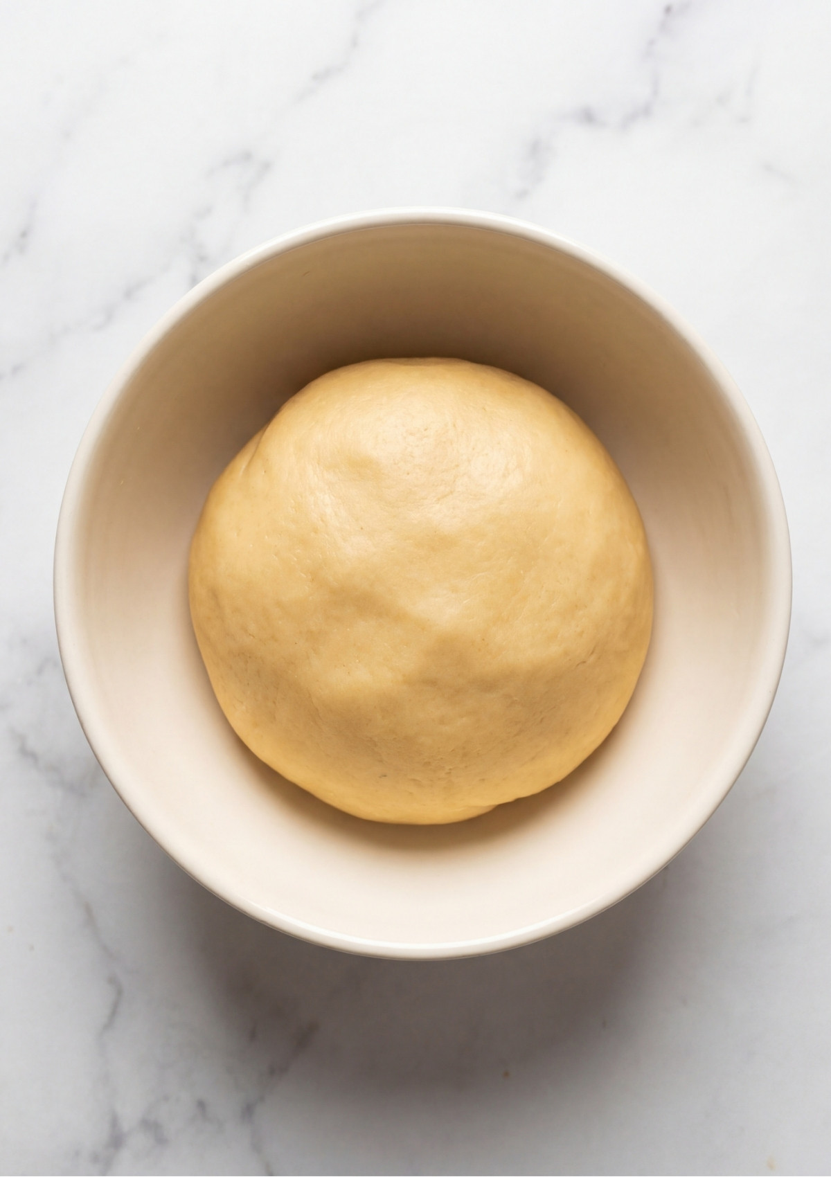 A smooth, pale yellow ball of chilled pastry dough resting in a beige ceramic bowl on a marble countertop, ready for a Patriotic Tartlets Recipe for Summer Parties.