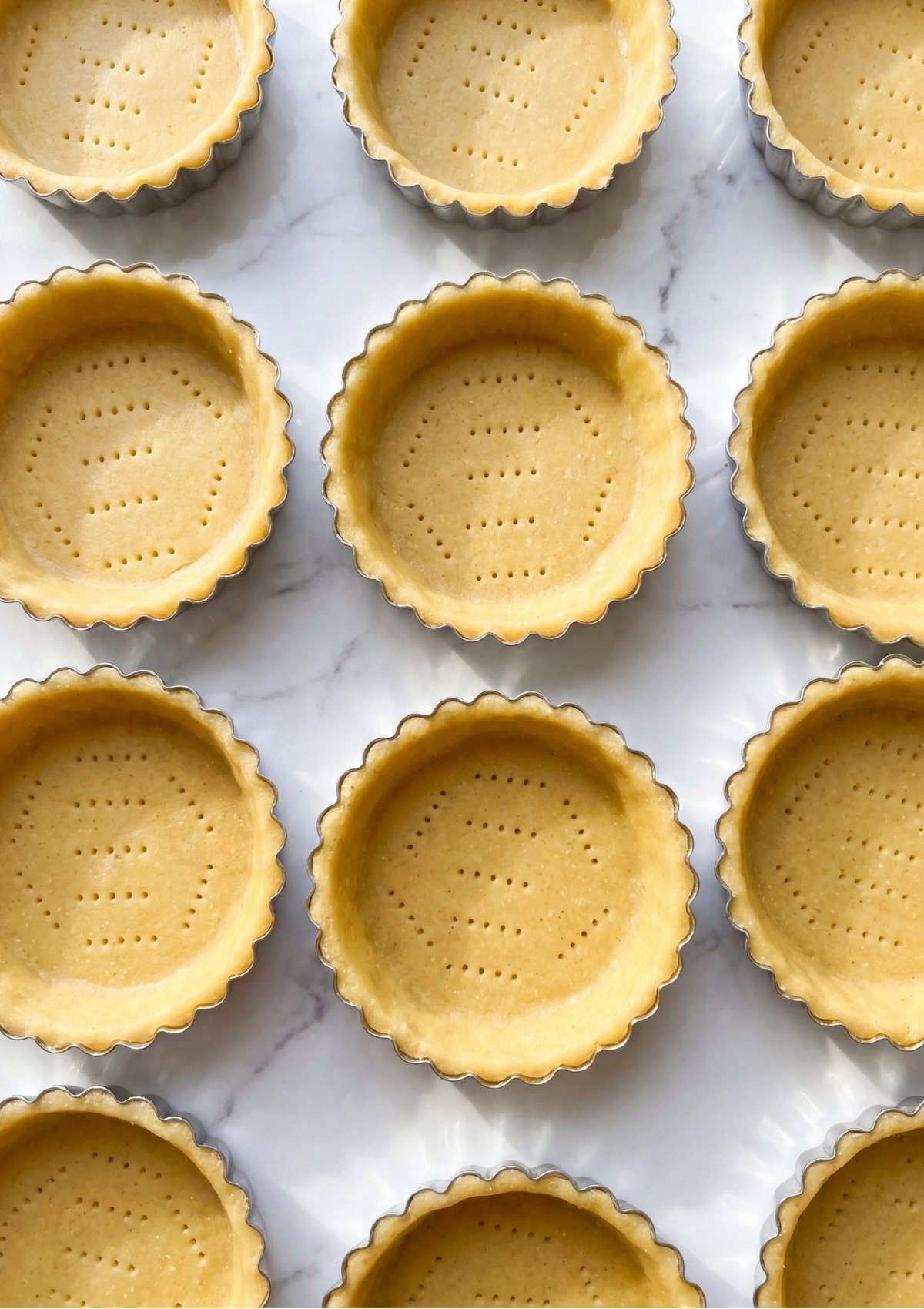 An overhead view of uncooked pastry dough pressed into fluted mini tart pans, with the bottoms pricked with a fork to prevent air bubbles during baking.