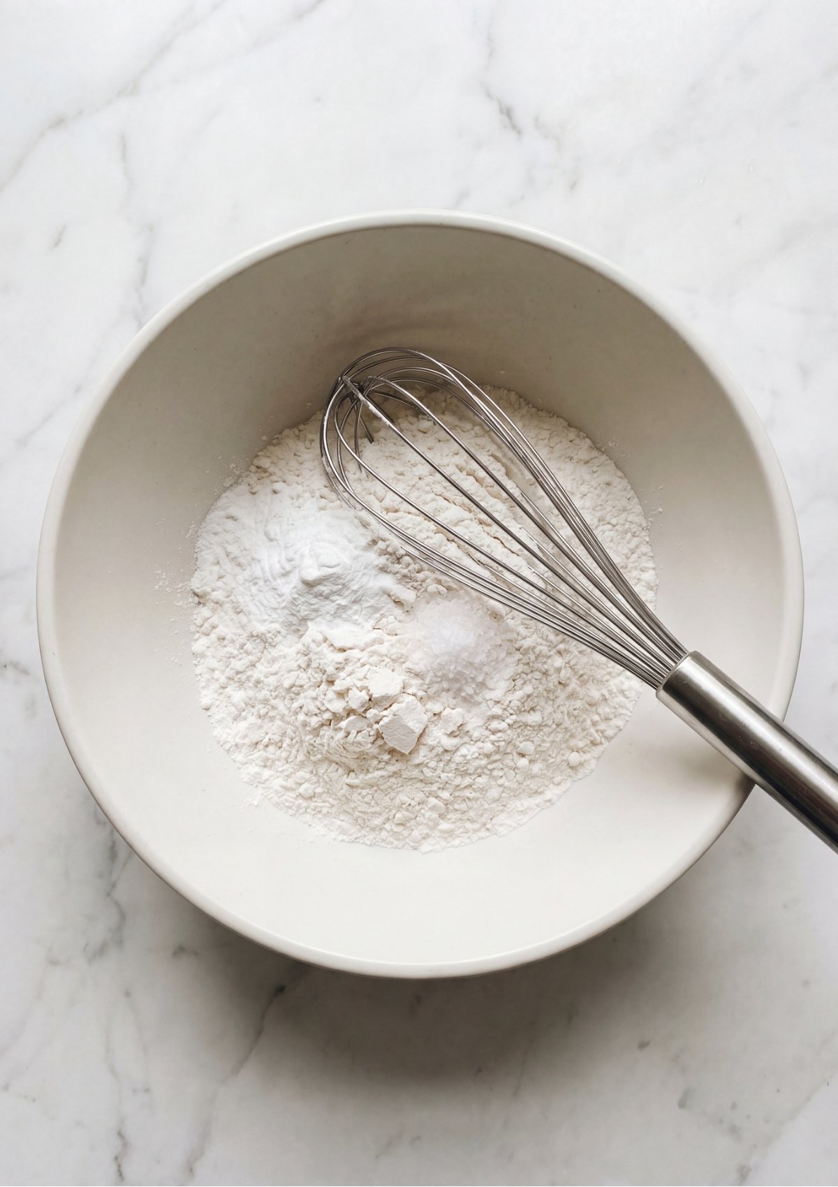 A whisk resting in a white bowl filled with all-purpose flour, baking powder, and a pinch of salt.