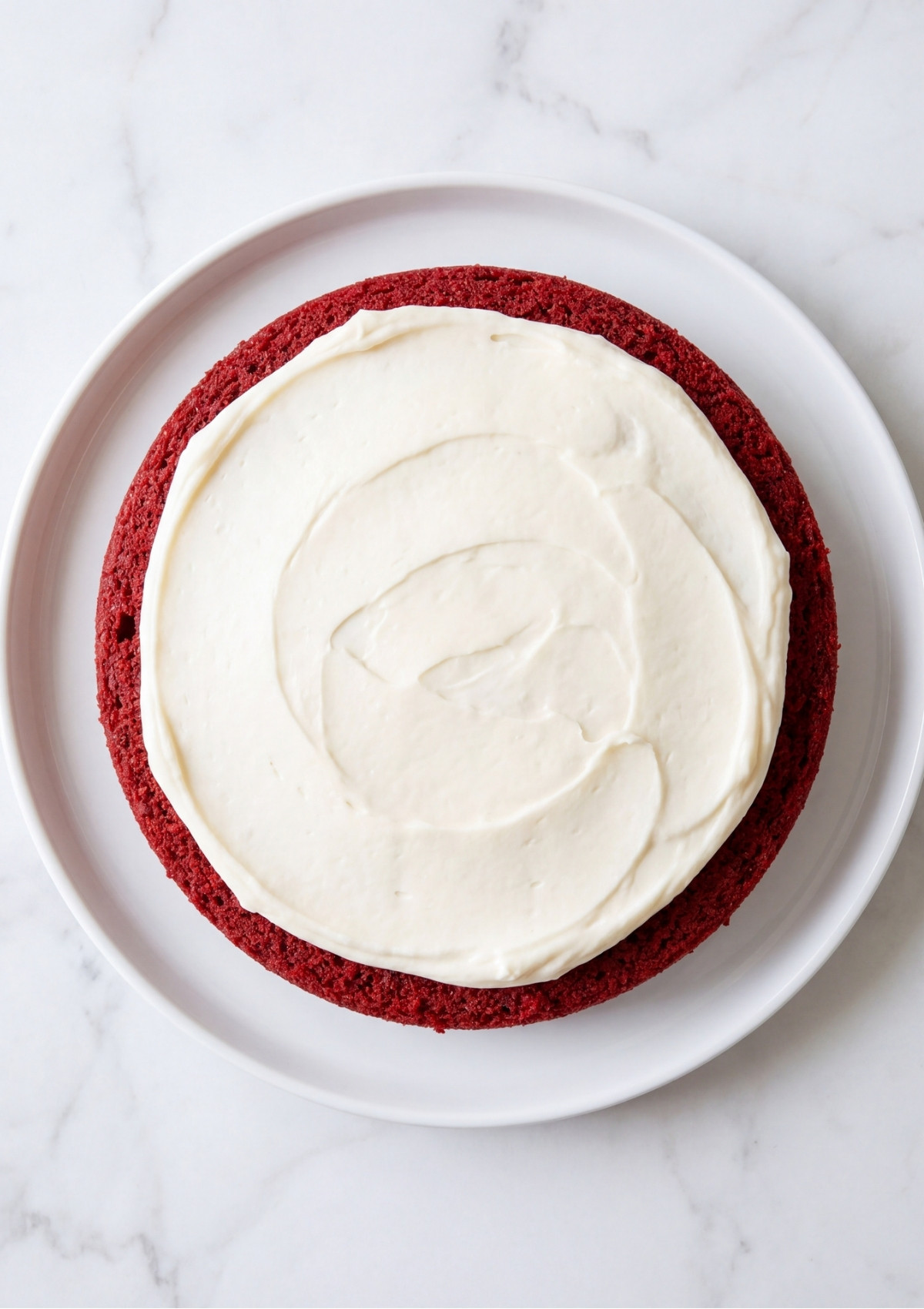 An overhead view of a round red cake layer topped with a smooth layer of white frosting on a white plate.