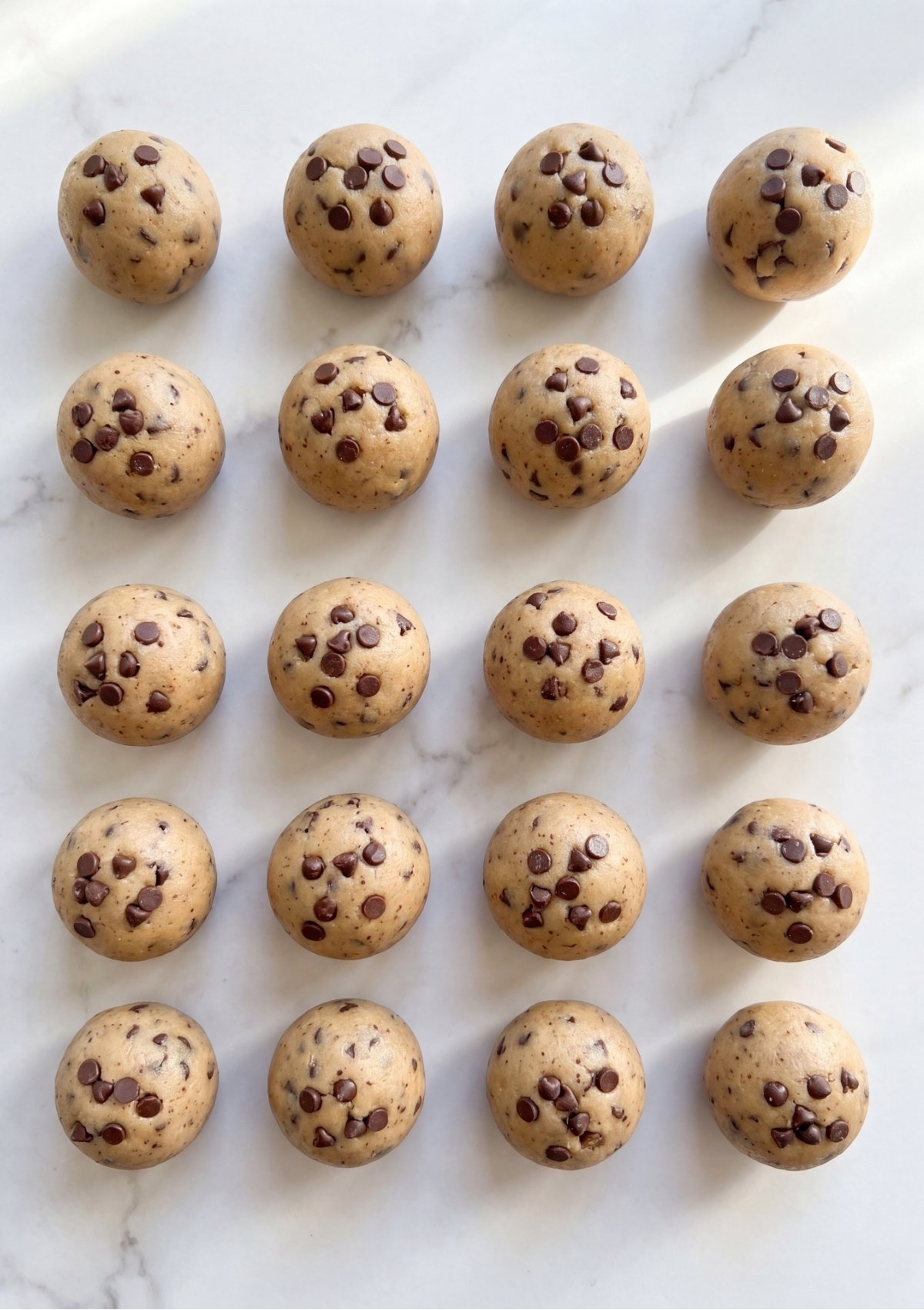 Multiple round edible chocolate chip cookie dough balls perfectly arranged in straight rows on a white marble surface