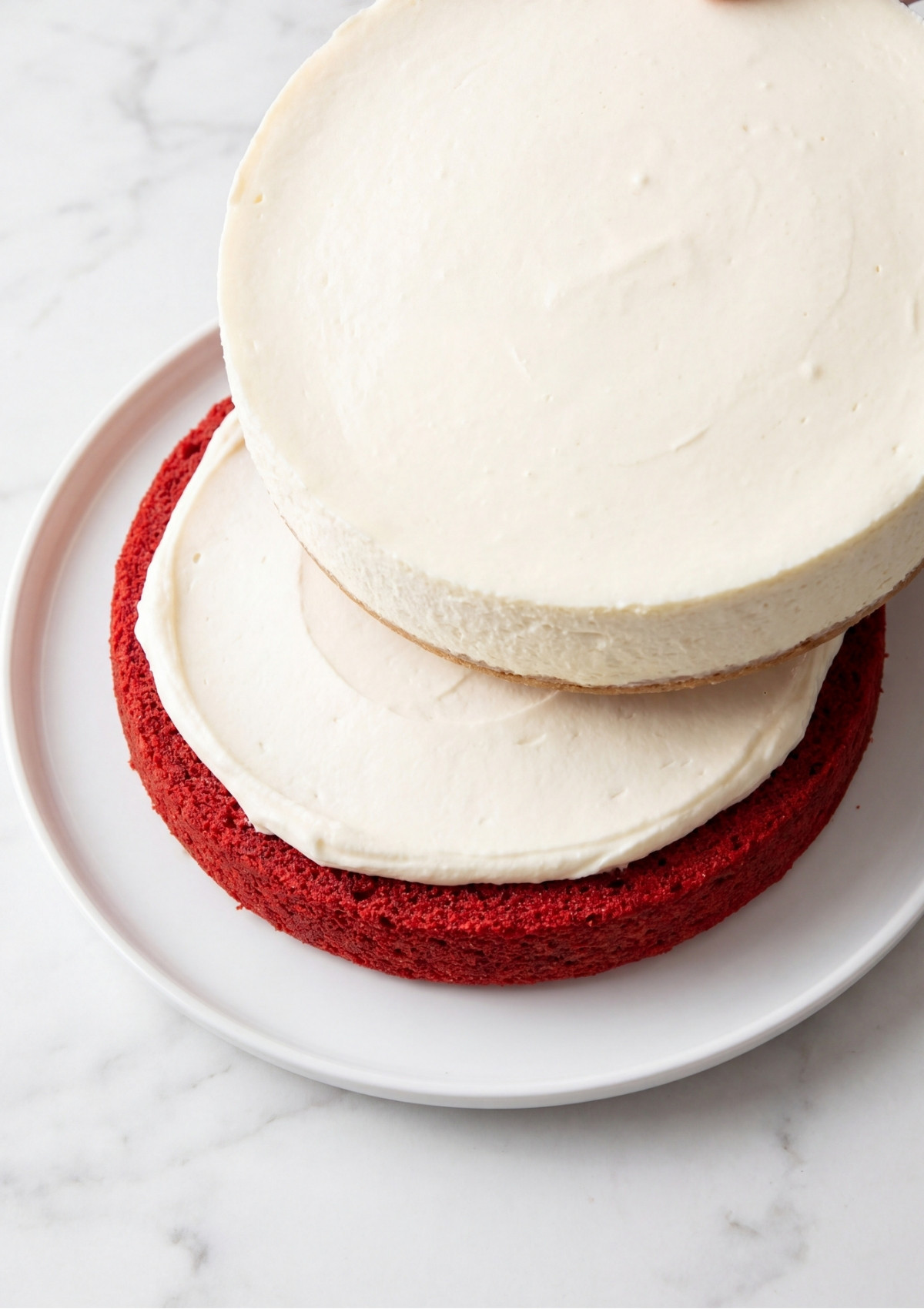 A cheesecake layer being placed on top of a red cake layer with white frosting on a white plate.