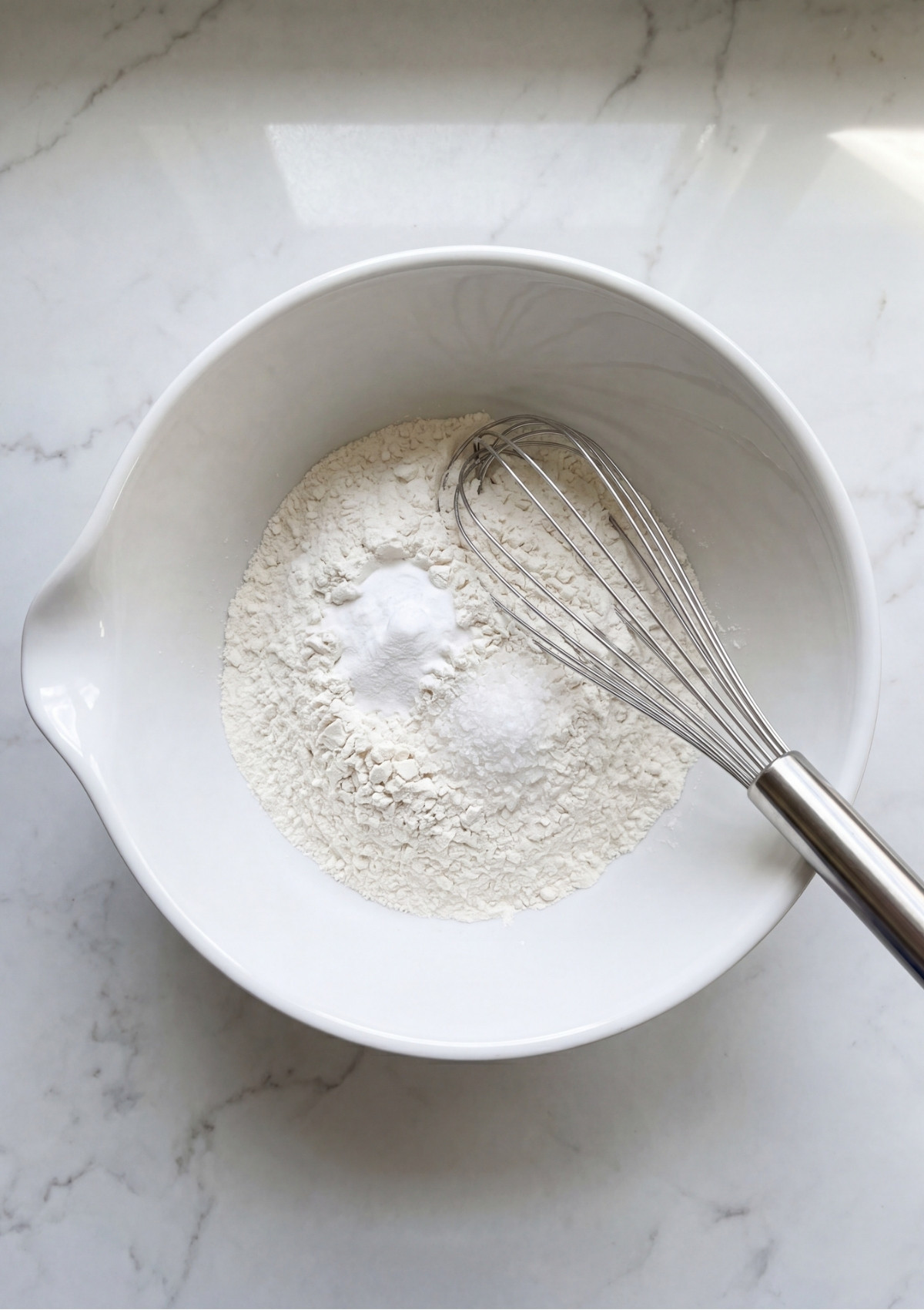 A white mixing bowl containing flour, baking powder, and salt with a metal whisk resting inside.