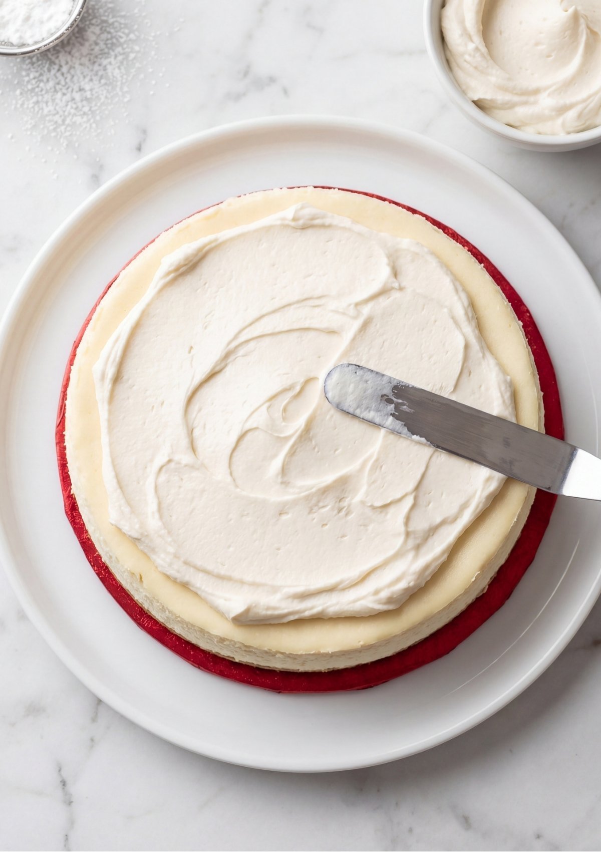 An overhead view of a metal spatula spreading white frosting over a cheesecake layer for a 4th of July cake.
