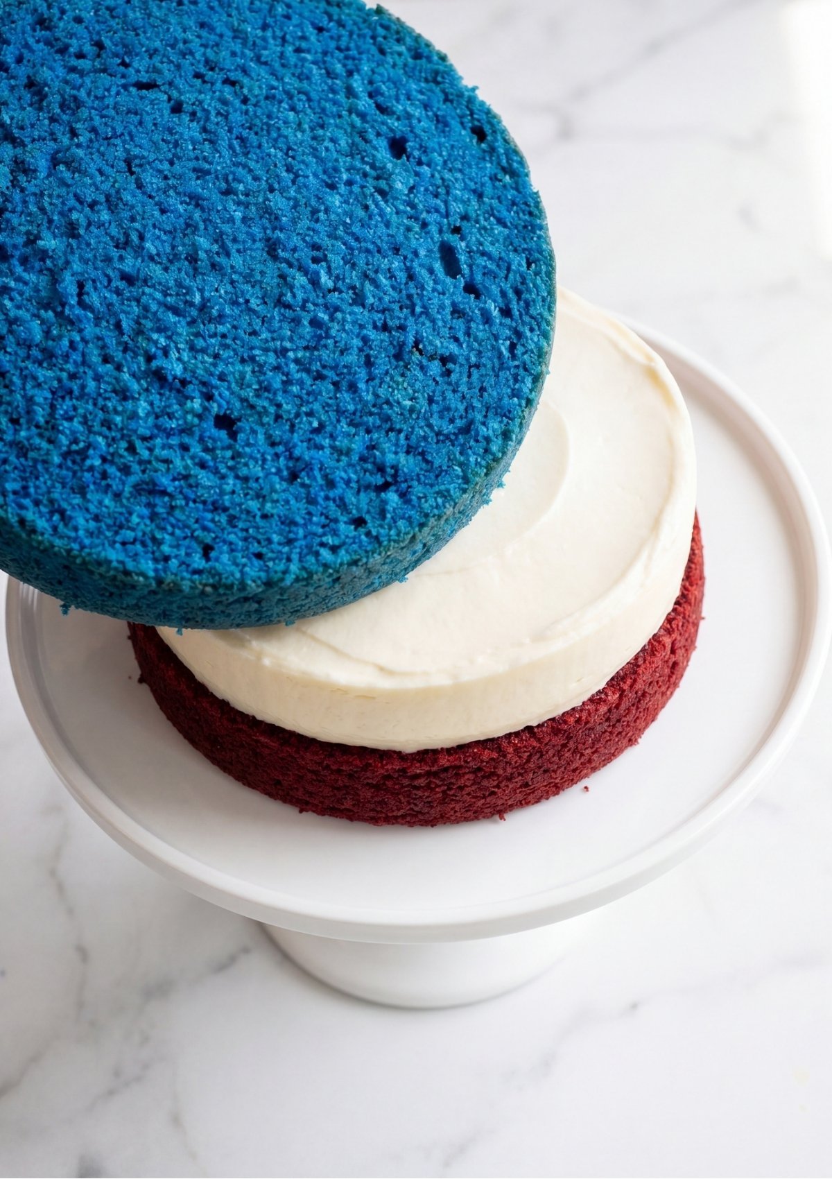 A blue cake layer being placed on top of a red cake and cheesecake stack with white frosting on a white stand.