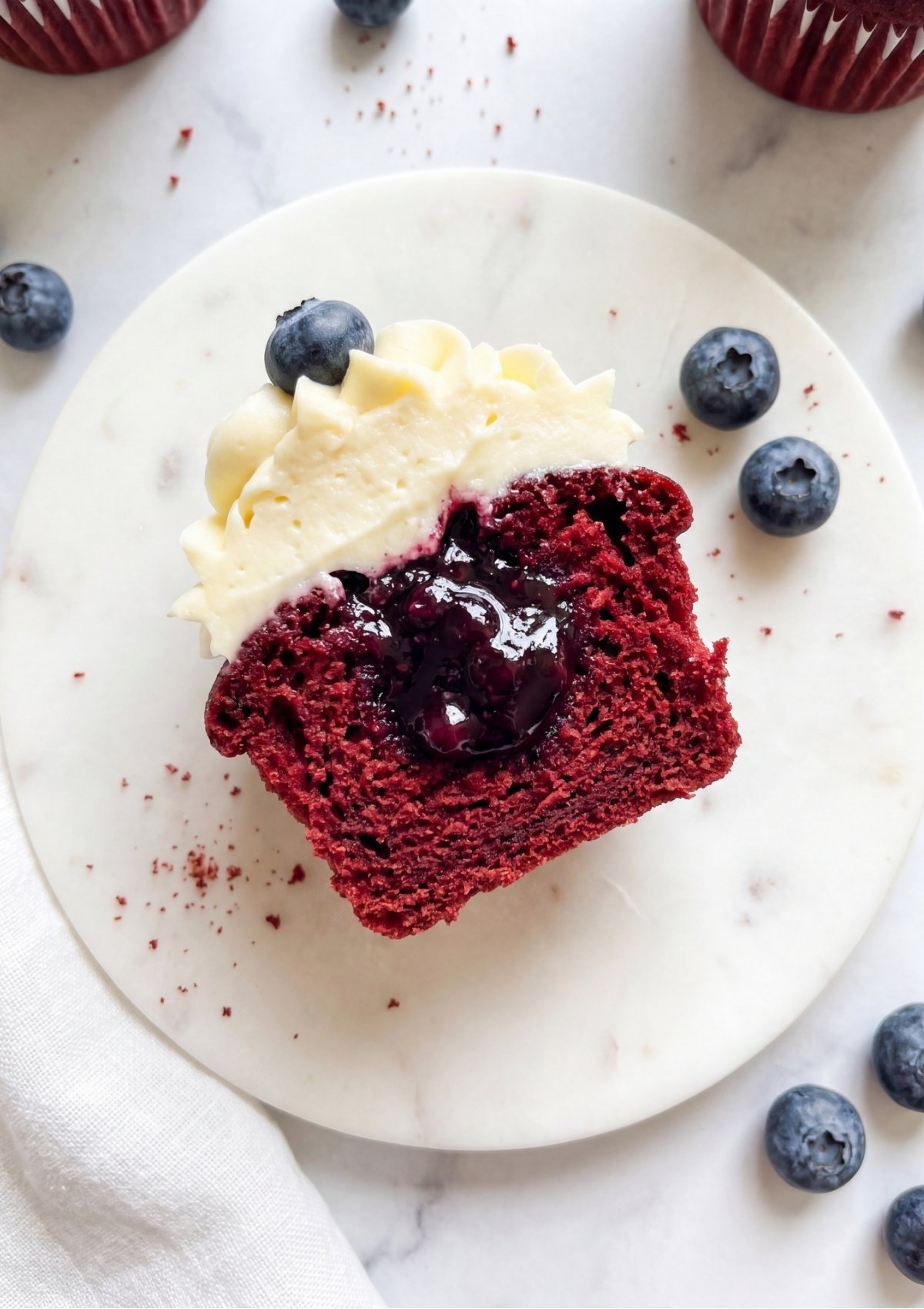 A red velvet cupcake sliced in half on a white marble coaster, revealing a hidden center filled with thick, glossy blueberry compote.