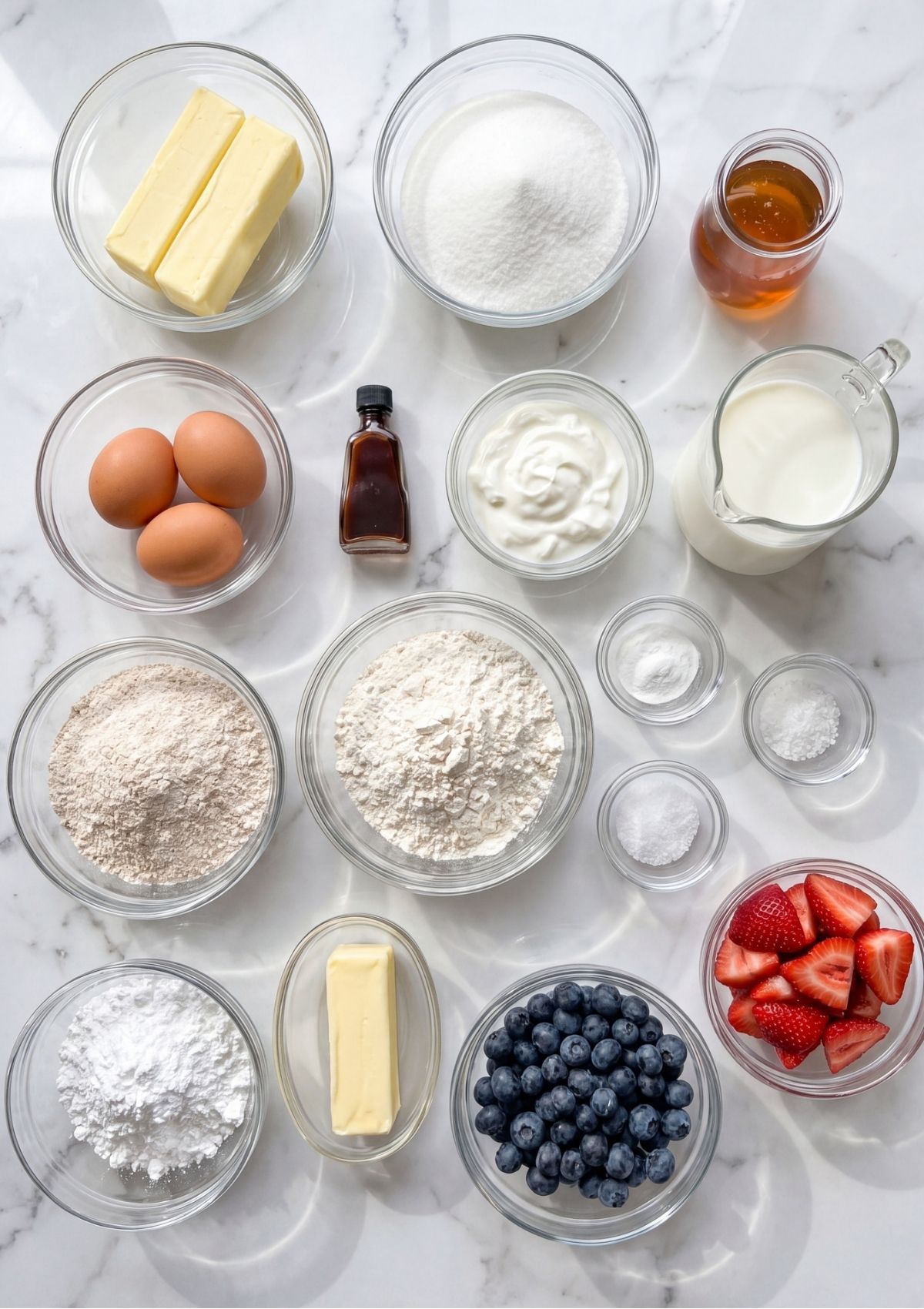 An overhead view of various bowls containing flour, sugar, eggs, butter, cream cheese, blueberries, and strawberries on a white marble surface.