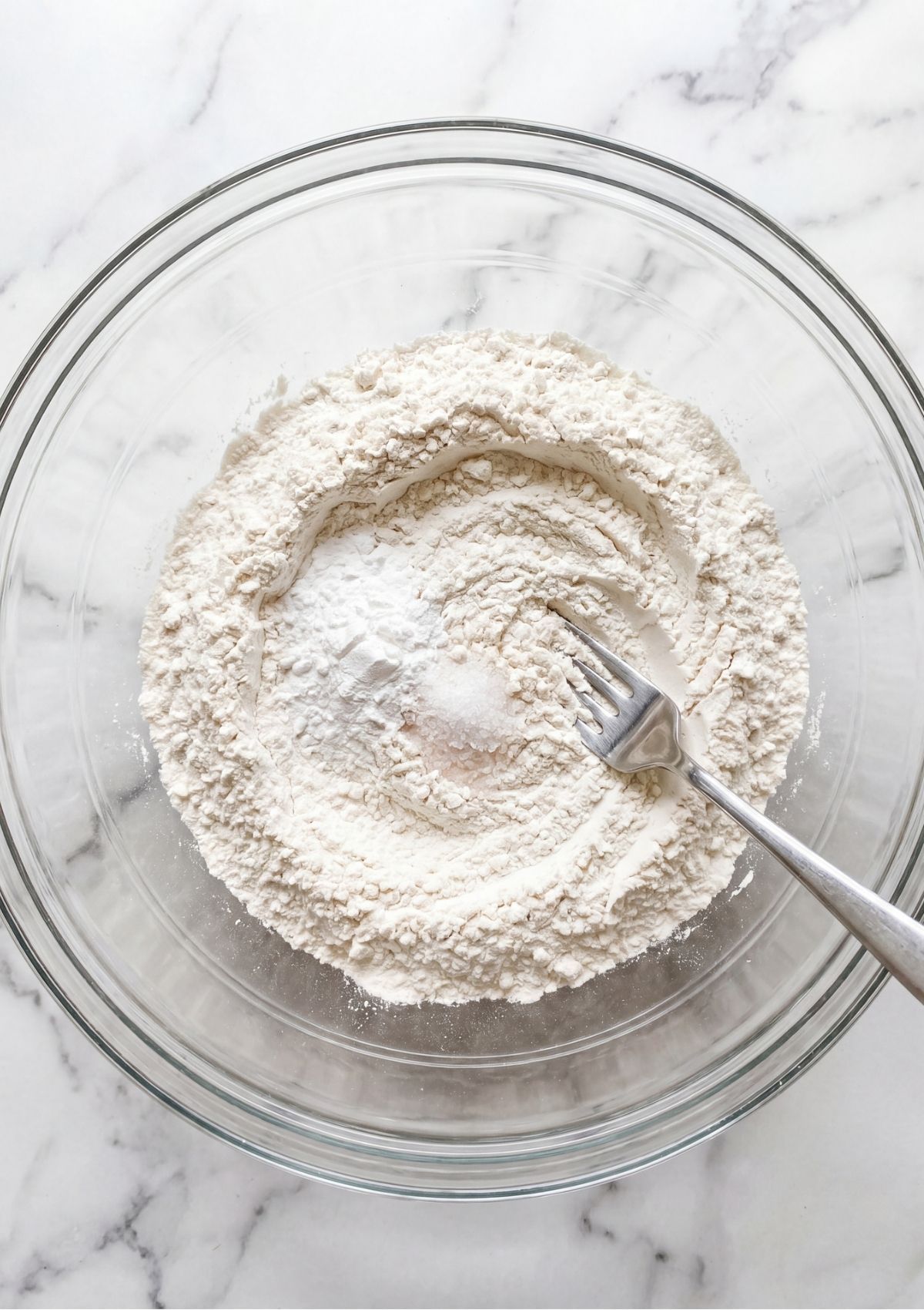 A glass bowl filled with flour baking powder and salt being mixed with a silver fork