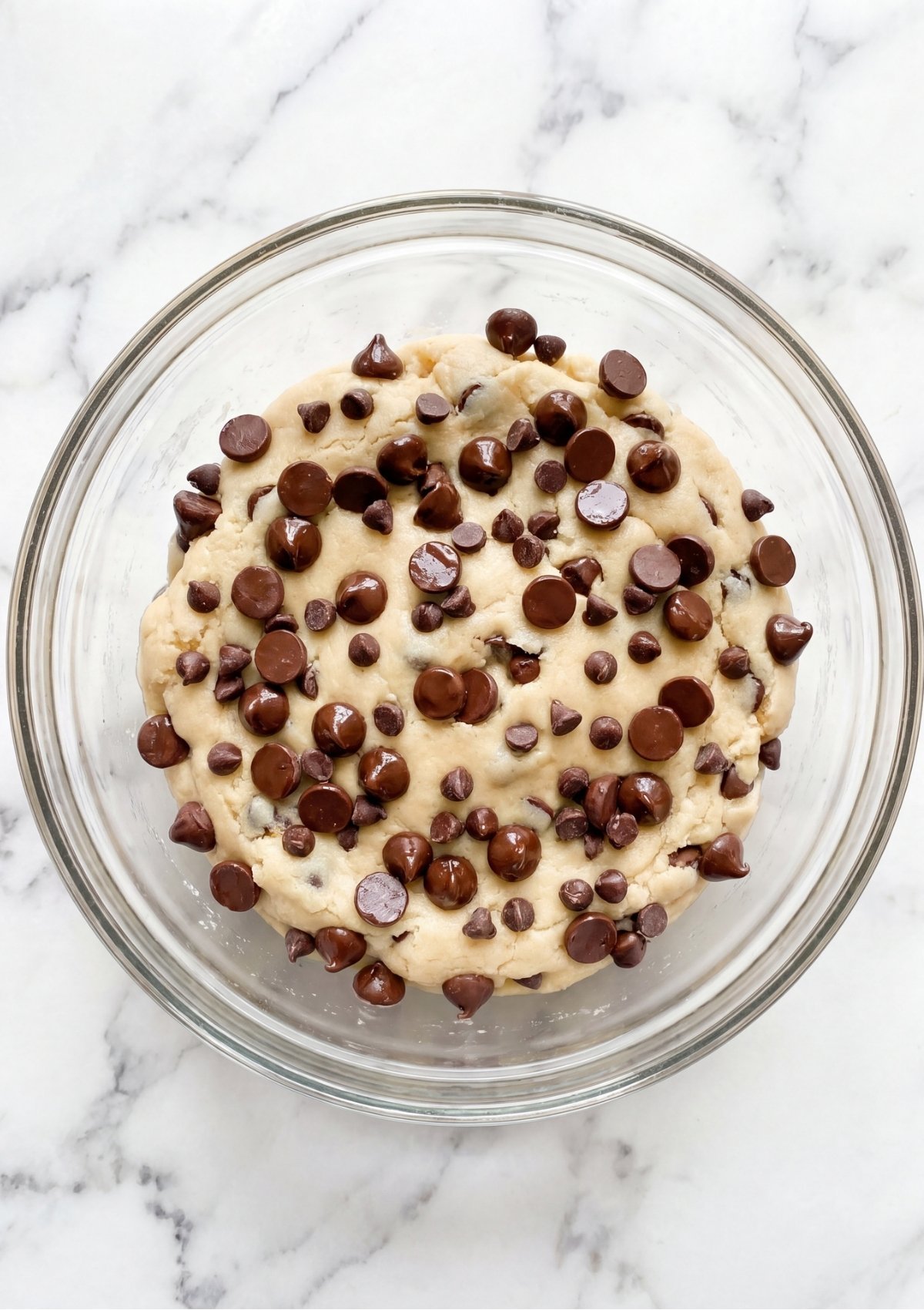 A glass mixing bowl filled with raw chocolate chip cookie dough topped with extra chocolate chips on a marble countertop
