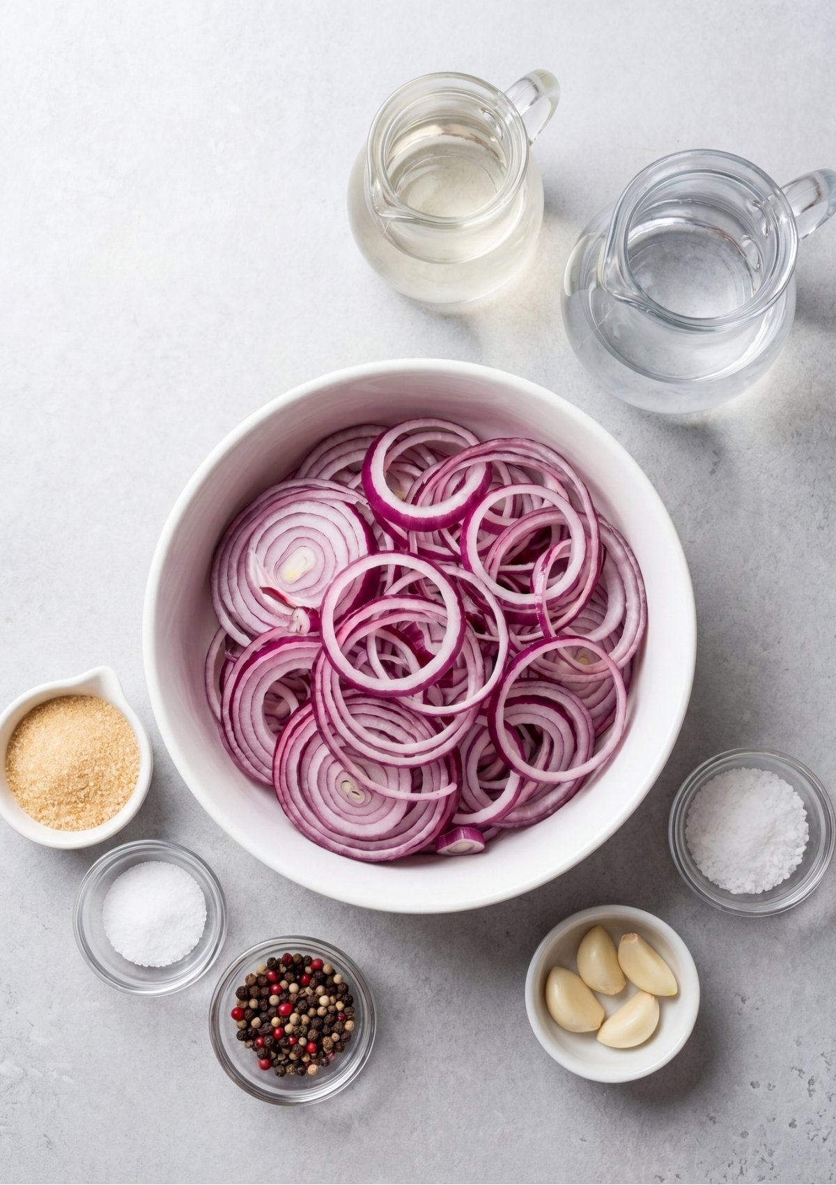 Sliced red onion rings in a white bowl surrounded by small glass dishes of sugar, salt, peppercorns, garlic, white vinegar, and water.