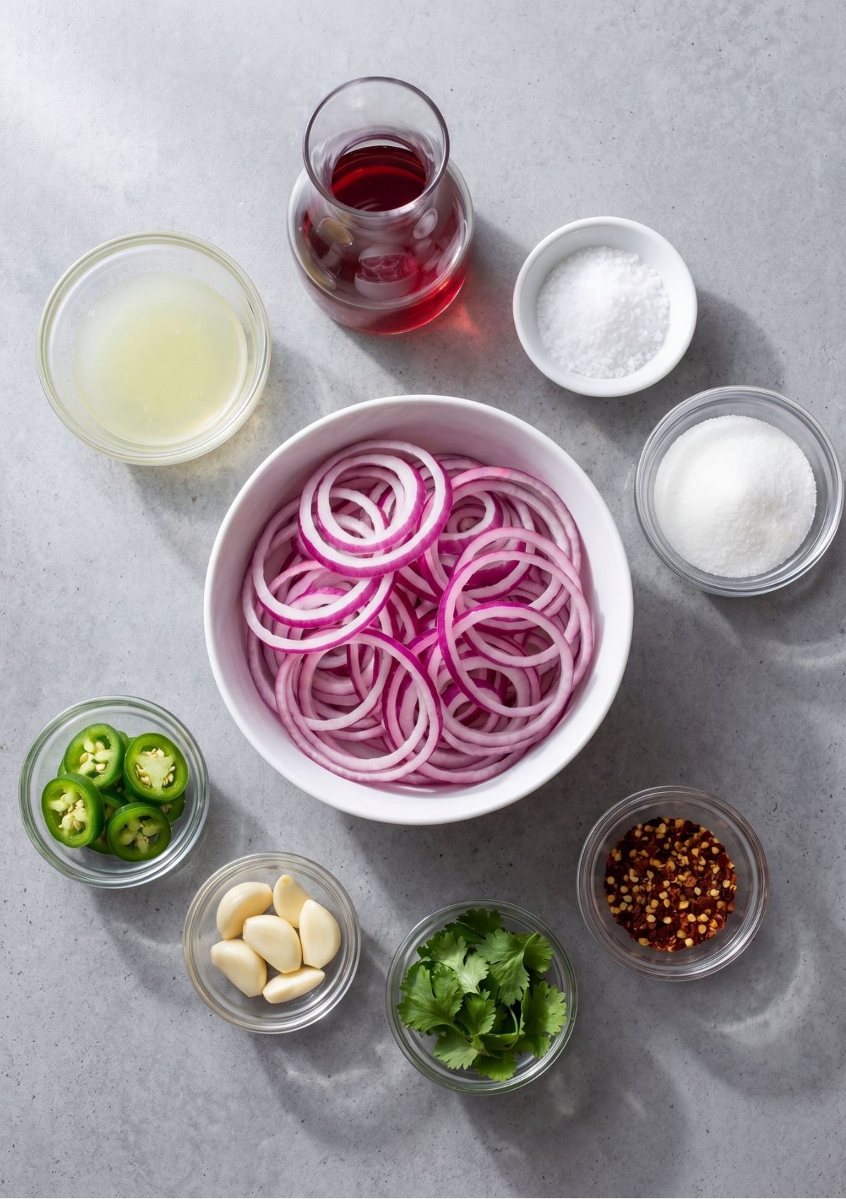 Prepped ingredients for Mexican pickled red onions arranged in separate small glass bowls around a larger white bowl of sliced red onion rings on a grey countertop
