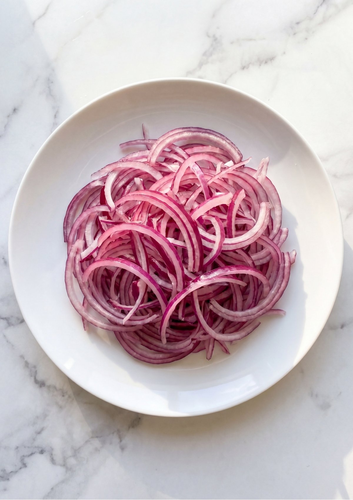 Thinly sliced raw red onions piled neatly in the center of a white round plate sitting on a marble countertop.