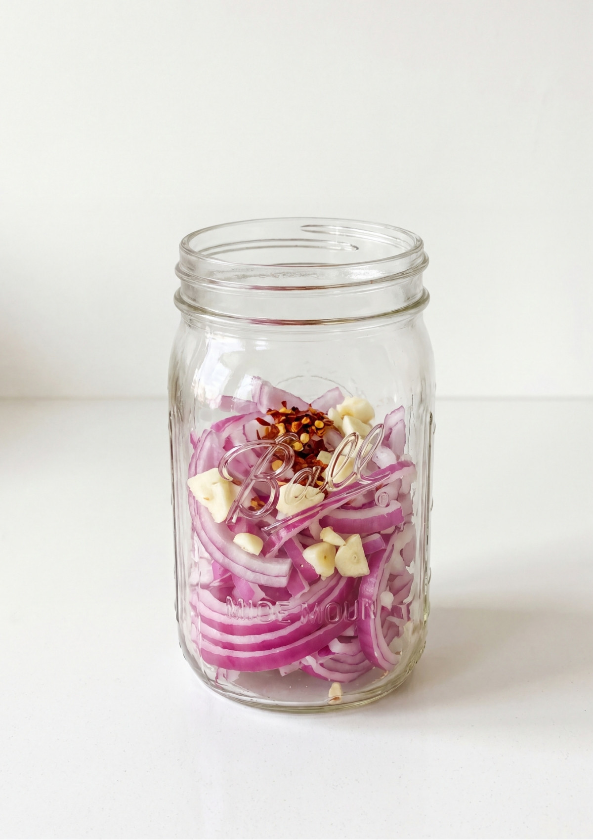 Sliced red onions, chopped garlic, and red pepper flakes layered at the bottom of an open glass mason jar