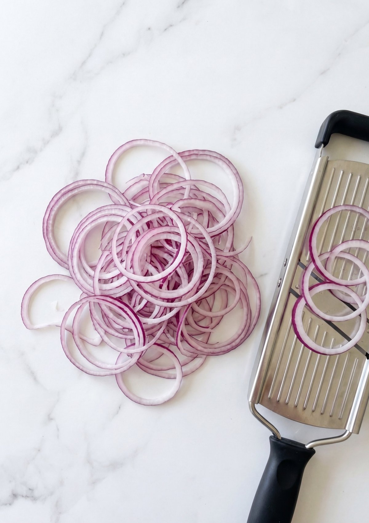 Top down view of a pile of thinly sliced red onion rings next to a metal mandolin slicer on a white marble countertop.