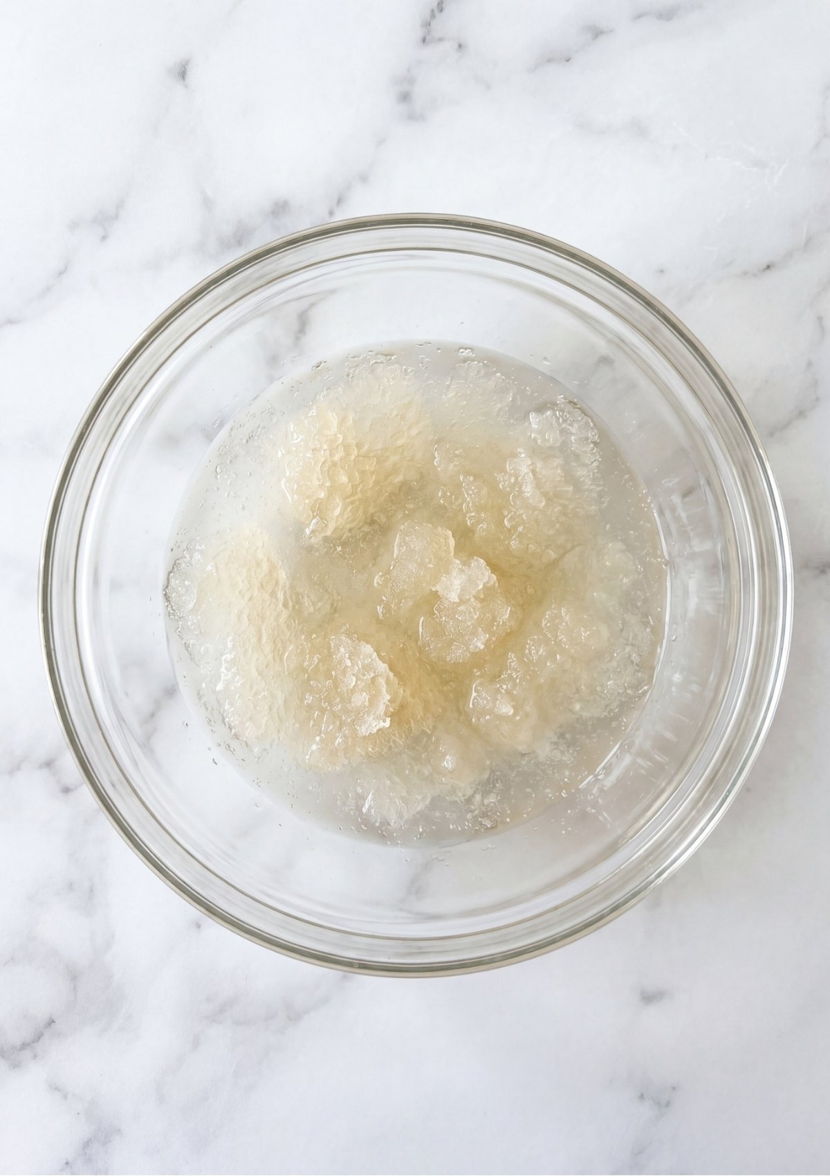 Unflavored gelatin powder blooming and thickening in cold water inside a clear glass bowl on a marble surface