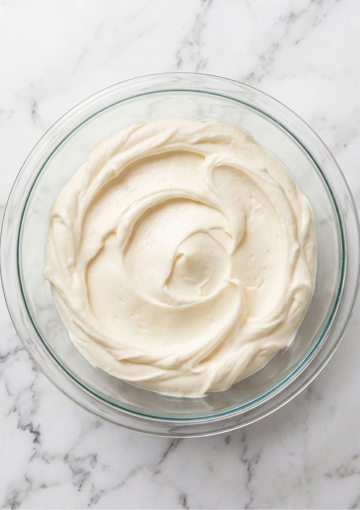 A clear glass bowl filled with smooth, whipped white cream cheese frosting, showing thick swirls ready for piping.