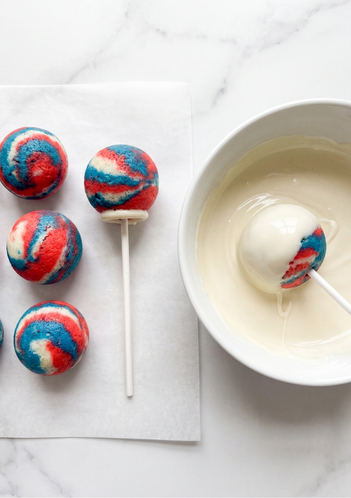 A hand-dipping a marbled cake pop into a bowl of smooth, melted white chocolate coating.