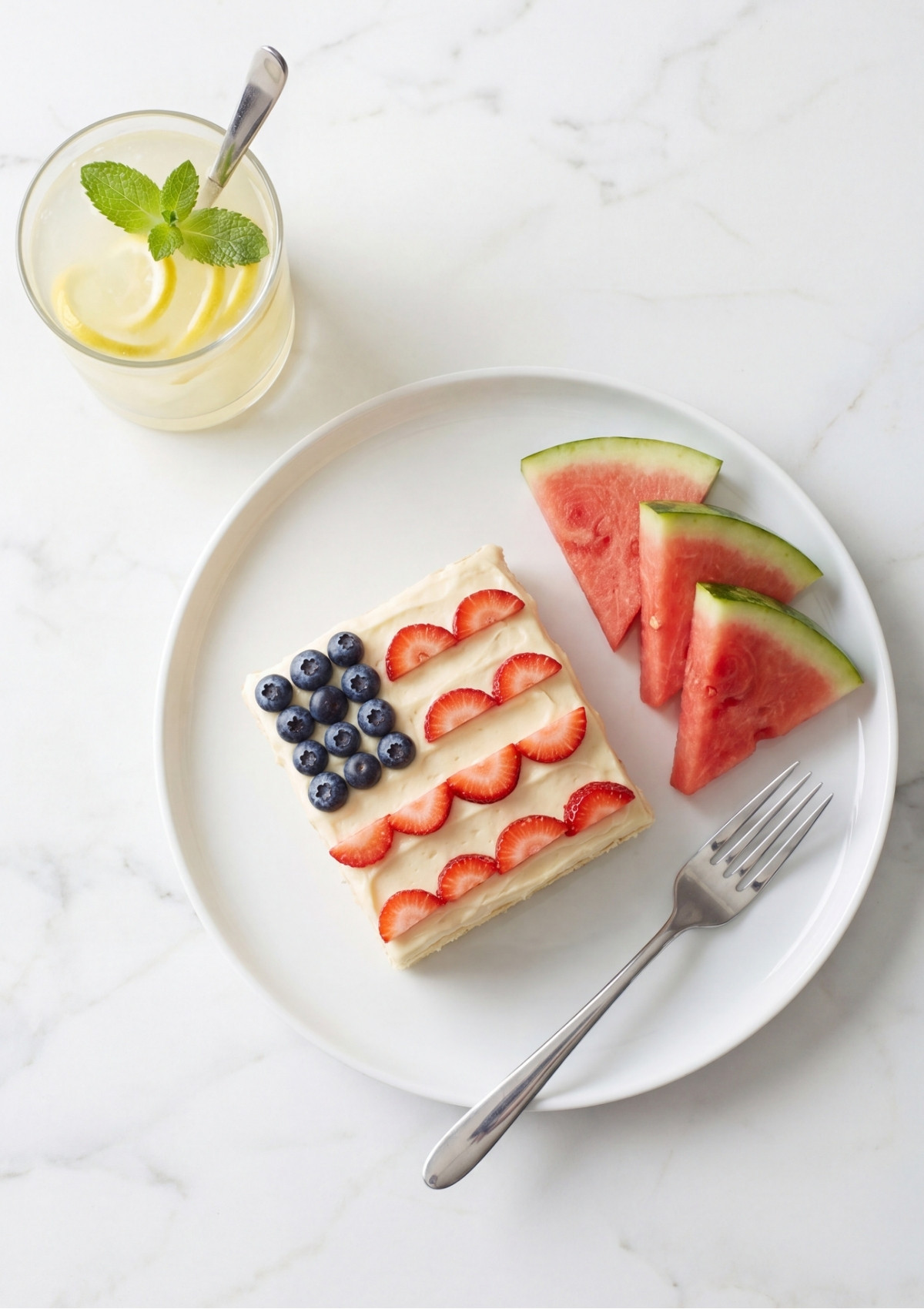 A white plate holding a slice of flag cake and three wedges of watermelon next to a glass of lemonade.