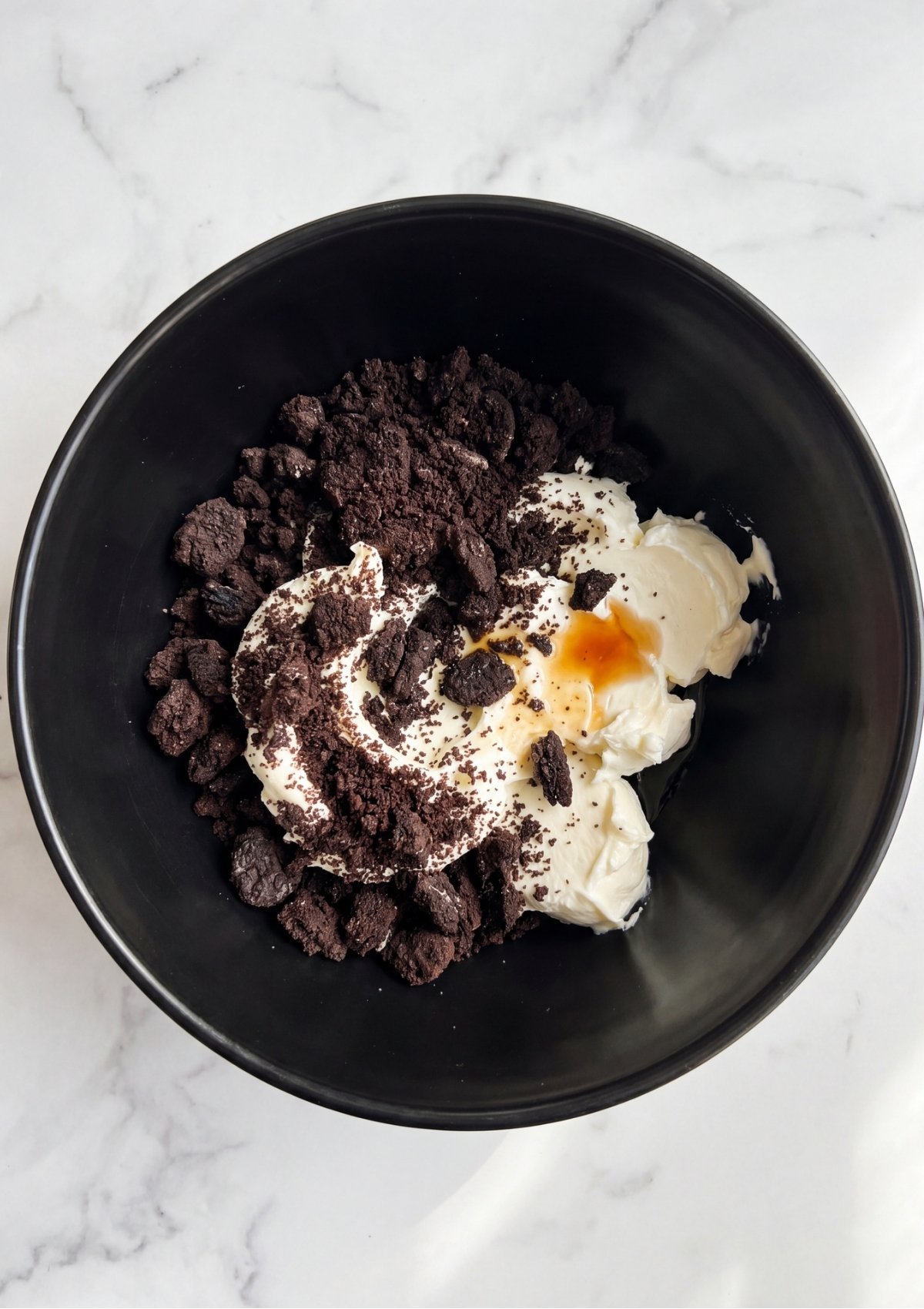 Crushed chocolate sandwich cookies being mixed with softened cream cheese and vanilla in a black bowl.