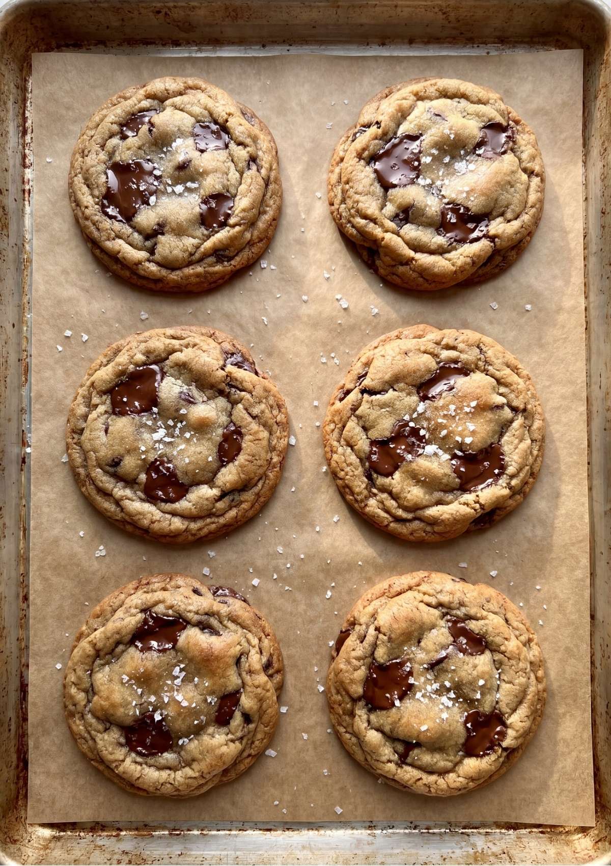 Six freshly baked chocolate chunk cookies with melted chocolate pools and flaky sea salt arranged on a parchment-lined baking sheet