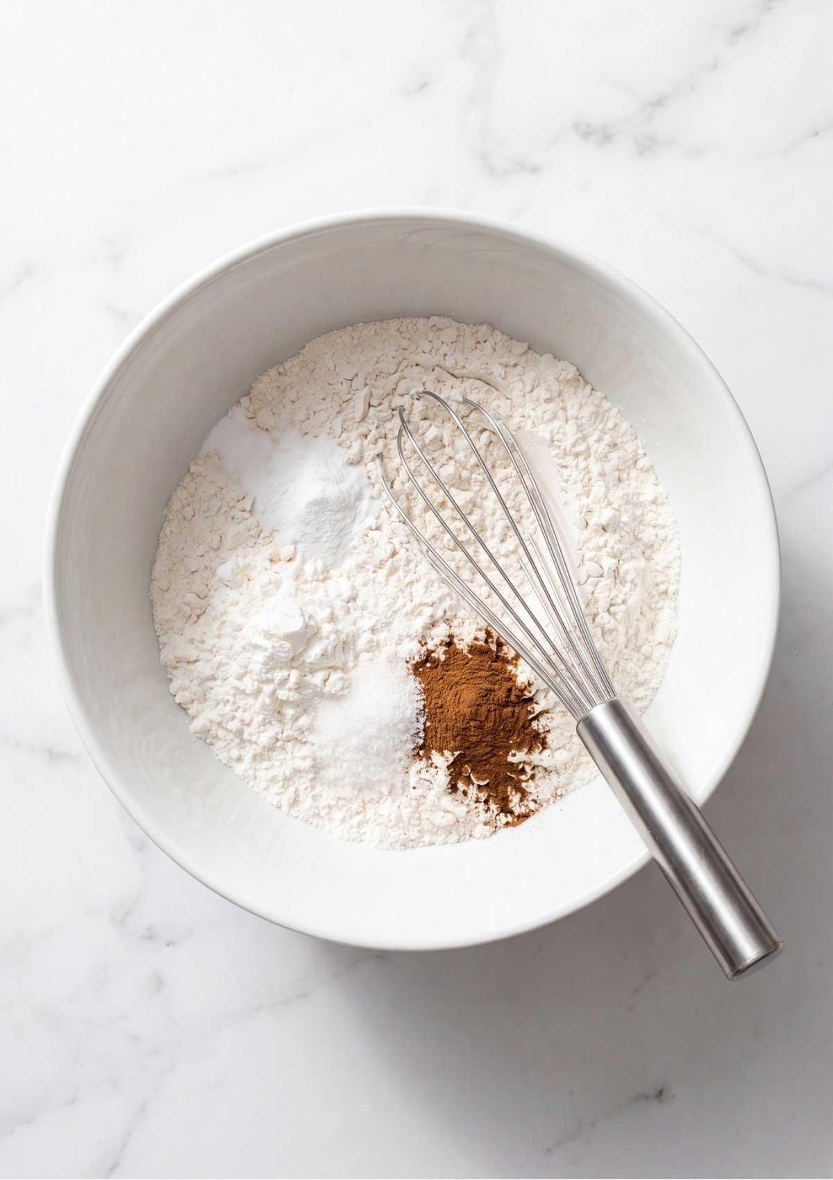 A white mixing bowl containing flour, cinnamon, baking soda, and salt with a metal whisk resting inside.